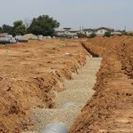 Obra de drenagem em área suburbana, com valas abertas e grandes tubulações instaladas no terreno.