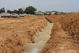 Obra de drenagem em área suburbana, com valas abertas e grandes tubulações instaladas no terreno.