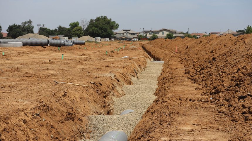 Obra de drenagem em área suburbana, com valas abertas e grandes tubulações instaladas no terreno.