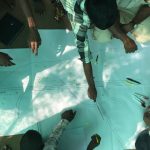 Grupo reunido ao ar livre analisa desenhos e ferramentas para planejar o traçado de uma via.