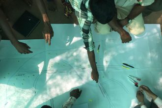 Grupo reunido ao ar livre analisa desenhos e ferramentas para planejar o traçado de uma via.