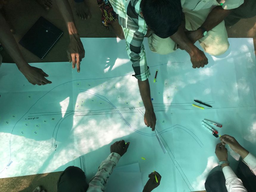 Grupo reunido ao ar livre analisa desenhos e ferramentas para planejar o traçado de uma via.