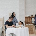 Criança conversa à mesa com adultos e uma profissional da assistência social em um ambiente doméstico.