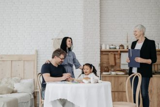 Criança conversa à mesa com adultos e uma profissional da assistência social em um ambiente doméstico.