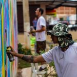 Dois artistas pintam um mural colorido em uma parede pública ao ar livre no Brasil.