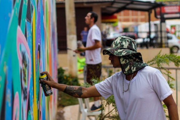 Dois artistas pintam um mural colorido em uma parede pública ao ar livre no Brasil.
