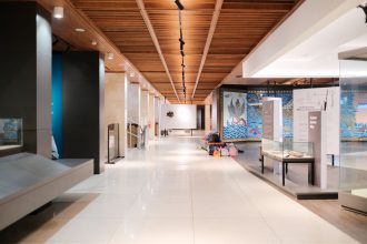Corredor iluminado de uma exposição de design moderno, com ambientes expositivos e passarelas vazias.