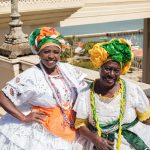 Duas mulheres negras sorriem ao ar livre usando trajes tradicionais baianos coloridos.