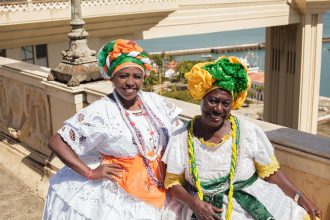 Duas mulheres negras sorriem ao ar livre usando trajes tradicionais baianos coloridos.