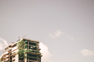 Edifício alto em construção, com estrutura aparente, contra o céu claro em uma área urbana.